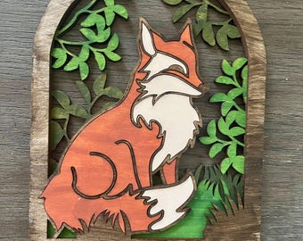 Op de afbeelding: Houten sleutelhanger met een vos-silhouet in een bos scène. De vos is oranje en wit met groene bladeren erachter. Er zijn vijf zwarte metalen haken aan de onderkant.