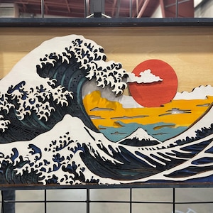 Puede incluir: Una pieza de arte de pared de madera que representa la Gran Ola de Kanagawa de Katsushika Hokusai. La ola es blanca con detalles azules y negros, y el fondo presenta un sol rojo, nubes amarillas y una montaña azul.