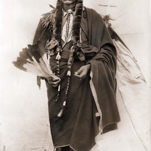 May include: A sepia photograph of a Native American man in traditional dress. He is wearing a dark robe, a beaded necklace, and holding a feathered fan. The image evokes a sense of cultural heritage and historical context.
