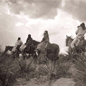 May include: A black and white photograph of four people riding horses through a field of tall grass. The people are wearing traditional clothing and the horses are moving forward. The sky is cloudy and the sun is shining.
