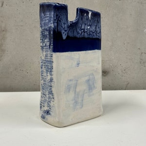 May include: A tall, rectangular ceramic vase with a unique, abstract design. The vase is glazed in shades of blue and white, with a textured surface. The top of the vase has a wide opening, making it suitable for displaying large flower arrangements.