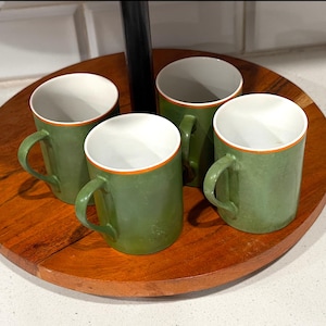 May include: A set of four cylindrical mugs with a matte olive green exterior and a white interior. Each mug features a thin, vibrant orange rim along the top edge and a matching green handle on the side, arranged on a circular wooden tray.