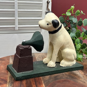 Puede incluir: Una figura de hierro fundido de un perro sentado de color crema con un collar negro, colocada junto a una bocina de fonógrafo de color marrón oscuro sobre una base rectangular verde. El perro mira hacia la bocina montada en un pedestal.