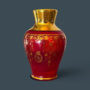 May include: A ruby red glass vase featuring a metallic gold neck and base. The body is decorated with intricate gold filigree patterns, floral motifs, and a circular emblem containing a winged lion figure and the letters VP in a stylized script.