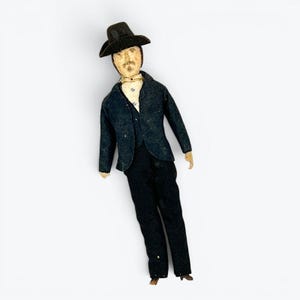 May include: A fabric doll featuring a figure wearing a dark charcoal gray suit, a matching wide-brimmed hat, and a light-colored shirt with small blue embroidered details. The doll has a painted face with a mustache and is dressed in formal attire.