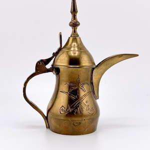 May include: A brass dallah coffee pot featuring a long, curved spout, a hinged lid with a pointed finial, and a curved handle. The body is engraved with a palm tree and two crossed swords, showcasing a metallic gold finish and a traditional silhouette.