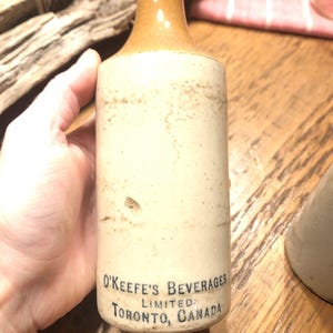 May include: A vintage ceramic bottle with a brown top and white body. The bottle has the text "O'Keefe's Beverages Limited Toronto, Canada" printed on the bottom.