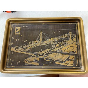 May include: A gold-toned metal tray with a dark brown background featuring a detailed engraved image of the 1939 New York World's Fair. The image includes the Trylon and Perisphere, the fair's iconic symbols.