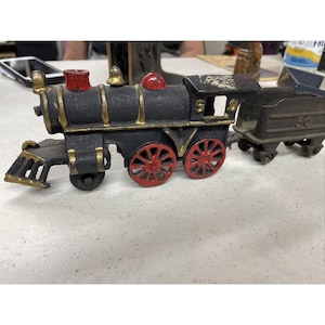 Puede incluir: Un tren de juguete de hierro fundido negro con ruedas rojas y detalles dorados. El tren tiene un vagón de carbón y una locomotora con una campana en la parte superior.