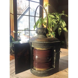 Puede incluir: Una antigua linterna de latón con una lente de vidrio rojo acanalado. La linterna tiene una puerta con bisagras y un asa para transportar. El metal tiene un aspecto envejecido. La linterna está abierta, revelando el cristal rojo. Un ventana y plantas son visibles en el fondo.