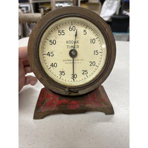 Op de afbeelding: Vintage Kodak Timer, een ronde, witgekleurde, metalen timer met zwarte markeringen en een zwarte wijzer. De timer is gemonteerd op een rode metalen basis. De timer wijzerplaat geeft "Kodak Timer" en "Eastman Kodak Co. Rochester, N.Y."