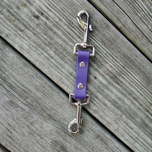 May include: A purple leather dog leash splitter with two silver-colored metal swivel snap hooks. The leash connector is on a weathered wooden surface.