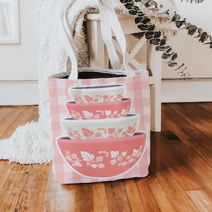 May include: A pink and white checkered tote bag featuring a stack of mixing bowls with floral designs. The bowls are in shades of pink and white. The bag has white handles and is sitting on a wooden floor.