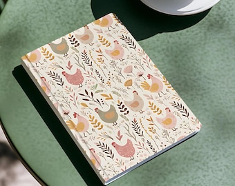 May include: A closed notebook with a cream-colored cover featuring a repeating pattern of chickens and foliage in shades of pink, yellow, and green. A white coffee cup and saucer with black coffee sit on a green table.