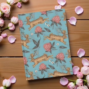 May include: A notebook with a teal background featuring a repeating pattern of running deer, pink clover flowers, and green leaves. The notebook is surrounded by pink rose petals and roses on a wooden surface.
