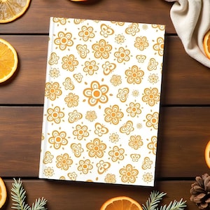 May include: A white notebook featuring a pattern of orange floral designs. The notebook is rectangular and rests on a wooden surface, accompanied by orange slices, pine cones, and foliage.