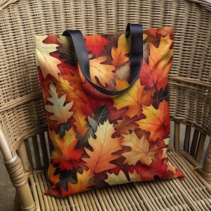 May include: A tote bag featuring a vibrant autumn leaf pattern in shades of red, orange, and yellow. The bag has black handles and is sitting on a wicker chair. The bag is designed for carrying items.
