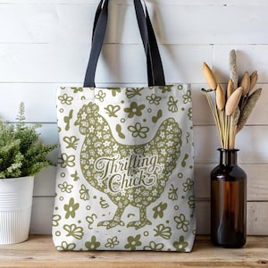 May include: White tote bag with black handles featuring a green floral chicken design and the text "Thrifting Chick." The bag is decorated with green floral patterns. The bag is sitting on a wooden surface.