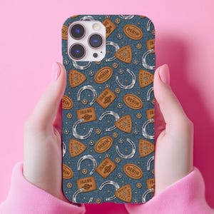 May include: A denim-patterned phone case featuring a repeating design of horseshoes, leather patches with the word "Denim", and circular accents. The case is held by a person with pink nails and a pink sweater.