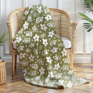 Pode incluir: Uma manta verde com um padrão floral branco drapeada sobre uma cadeira de vime. A manta apresenta um design repetido de flores brancas estilizadas sobre um fundo verde sólido. A cadeira está numa sala com uma planta.