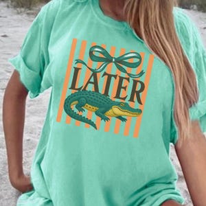 May include: A light teal t-shirt with a graphic featuring an alligator, the word "LATER", and orange stripes. The alligator is green and yellow. The shirt has rolled-up sleeves.