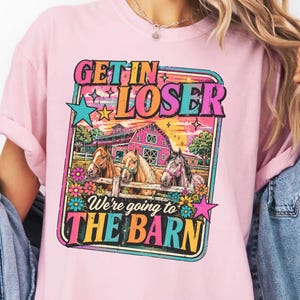 May include: Pink t-shirt with a colorful graphic that reads "GET IN LOSER" above an image of horses in front of a barn. The text "We're going to THE BARN" is below the image. The shirt has short sleeves.