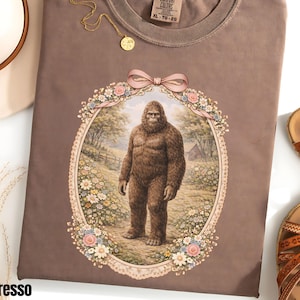 May include: Espresso-colored t-shirt with a Bigfoot graphic in a floral frame. The shirt has a crew neck and short sleeves. A gold necklace and a pair of sandals are also visible.