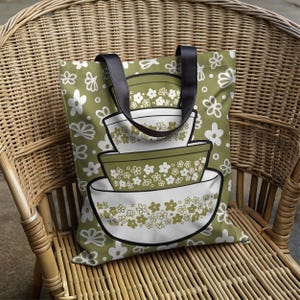 May include: A green tote bag with a floral pattern and black handles. The bag features a stack of white and green bowls with floral designs. The bag is sitting on a wicker chair.