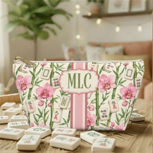 Può includere: Una pochette con cerniera su sfondo color crema con un motivo di orchidee rosa, steli di bambù verdi e tessere del mahjong. Una striscia verticale rosa centrale contiene una cornice decorativa con le lettere MLC in un carattere serif verde.