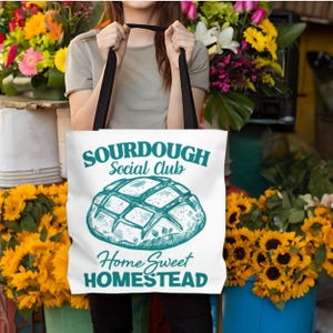Könnte beinhalten: Weiße Tragetasche mit schwarzen Henkeln. Auf der Tasche steht der Text "SOURDOUGH Social Club" und "Home Sweet HOMESTEAD" in Türkis, sowie eine türkisfarbene Illustration eines Brotlaibs.