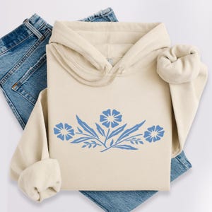 Könnte beinhalten: Cremefarbener Kapuzenpullover mit blauem Blumendesign und Kapuze. Der Kapuzenpullover wird mit blauen Jeans kombiniert. Das Blumendesign zeigt drei Blüten und Blätter. Die Ärmel des Kapuzenpullovers sind umgeschlagen.