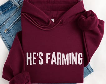 May include: A maroon hoodie with the text "HE'S FARMING" in white block letters. The hoodie is folded on top of a pair of blue denim jeans. The hoodie has a drawstring and a hood.