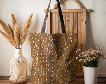 May include: Brown tote bag featuring a white spotted pattern, similar to deer fur. The bag has black handles and is shown with decorative items, including a vase with dried plants and a wooden frame.