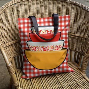 May include: A tote bag with a red and white checkered pattern and black handles. The bag features a graphic of four nested mixing bowls in red, white, and yellow, each with a decorative bird and floral design. The bag is sitting on a wicker chair.