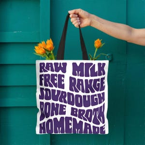 Könnte beinhalten: Weiße Tragetasche mit schwarzen Henkeln, mit den Worten "RAW MILK FREE RANGE SOURDOUGH BONE BROTH HOMEMADE" in lila, stilisierten Buchstaben. Die Tasche ist mit orangefarbenen Blumen gefüllt. Der Hintergrund ist eine türkisfarbene Tür.