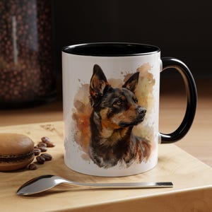 May include: A white ceramic mug with a black rim and handle features a watercolor painting of a brown and black Australian Shepherd dog. The dog is looking to the right and has a brown nose and brown eyes. The background is a watercolor wash of brown, orange, and yellow.