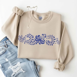 May include: A beige sweatshirt with a blue floral design across the chest. The design features stylized flowers and leaves. The sweatshirt is folded with a pair of blue jeans and a silver necklace.