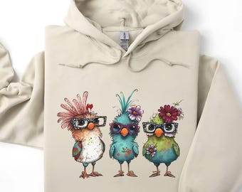 May include: Beige hoodie featuring a whimsical design of three colorful birds wearing glasses and floral accents. The birds are in shades of orange, blue, and green. The hoodie has a drawstring hood and a kangaroo pocket.