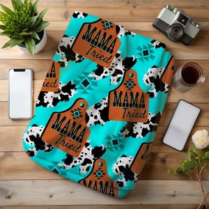 May include: A turquoise blanket with a cowhide pattern and brown tags that read "MAMA Tried". The blanket also features geometric designs in black and turquoise. A camera, coffee, and a smartphone are visible.