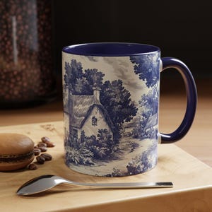May include: A ceramic mug with a deep blue interior and handle, featuring a white exterior decorated with a blue toile-style print of a rural cottage landscape with trees and foliage, set against a neutral background with a spoon and cookie.