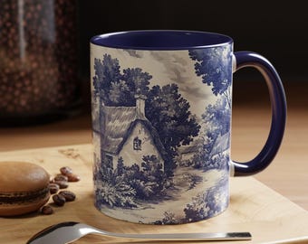 May include: A ceramic mug with a deep blue interior and handle, featuring a white exterior decorated with a blue toile-style print of a rural cottage landscape with trees and foliage, set against a neutral background with a spoon and cookie.