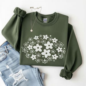Pode incluir: Uma camisola verde-oliva com um design floral branco. A camisola de gola redonda é combinada com jeans de ganga de lavagem clara e um colar de prata. O design floral é um aglomerado de flores brancas.