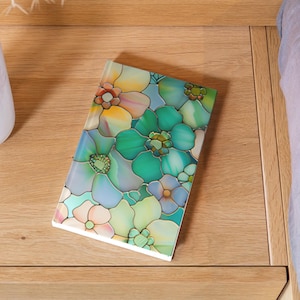 May include: A rectangular notebook with a floral design. The cover features a stained-glass style pattern of flowers in shades of green, blue, yellow, and pink. The notebook is resting on a wooden surface.