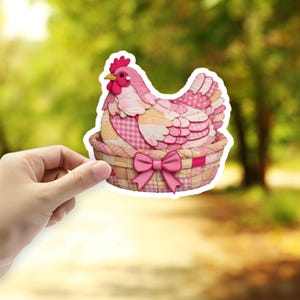 May include: A pink and white patchwork chicken sticker sitting in a woven basket with a pink bow. The chicken has a red comb and beak. The sticker is held up against a blurred green and yellow background.