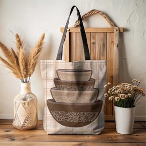 May include: A beige tote bag with a plaid pattern and black handles. The bag features a stack of four brown mixing bowls with white floral designs. The background includes a wooden frame, vase with pampas grass, and a small white pot with dried flowers.