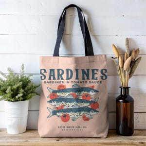May include: A light pink tote bag with black handles featuring a graphic of sardines and tomatoes. The text "SARDINES" is printed in blue, along with "SARDINES IN TOMATO SAUCE" and "EXTRA VIRGIN OLIVE OIL SARDINES CLUB".