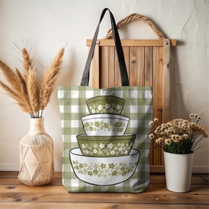 May include: A tote bag featuring a stack of four green and white floral patterned bowls. The bag has a green and white gingham pattern background. The bag has black handles and a brown leather strap. The bag is sitting on a wooden surface.