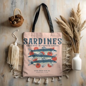 May include: A light pink tote bag with black handles featuring a graphic of sardines and tomatoes. The text "SARDINES" and "Sardines in Tomato Sauce" is printed in blue. The bag also includes the text "EXTRA VIRGIN OLIVE OIL SARDINES CLUB."