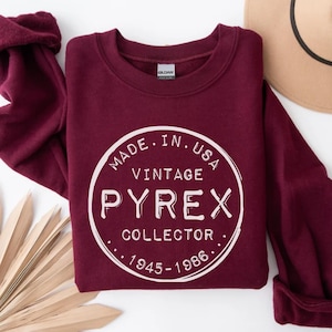 Op de afbeelding: Een bordeauxrode sweatshirt met een witte cirkelvormige afbeelding met de tekst "MADE IN USA VINTAGE PYREX COLLECTOR 1945-1986." De sweatshirt heeft een ronde hals en lange mouwen. Een beige hoed is gedeeltelijk zichtbaar.