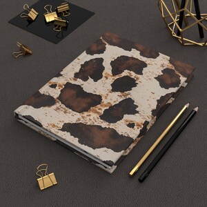 May include: A hardbound journal with a cowhide pattern in shades of brown and cream. Gold-colored binder clips and pencils are arranged around the journal on a dark gray surface. The journal is a stylish accessory for writing.